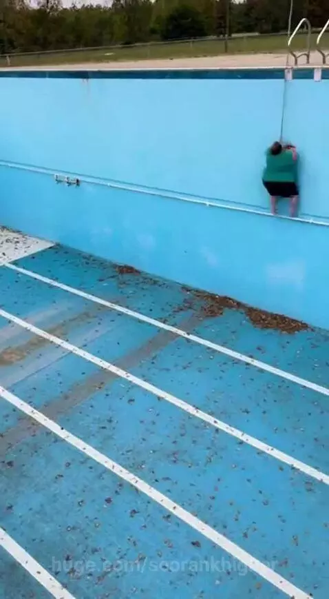 Woman in green shirt and black shorts falls onto the floor of an empty, debris-filled pool after a rope swing.