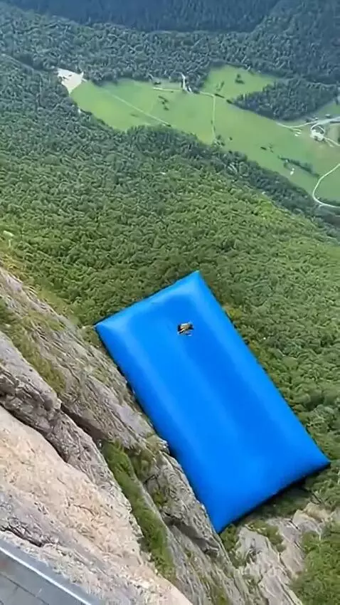 Woman in a red chair launching off a cliff, falling towards a blue inflatable mat below.