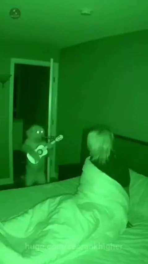 Night vision video shows a creature with glowing eyes playing a guitar in a doorway while a person lies in bed.