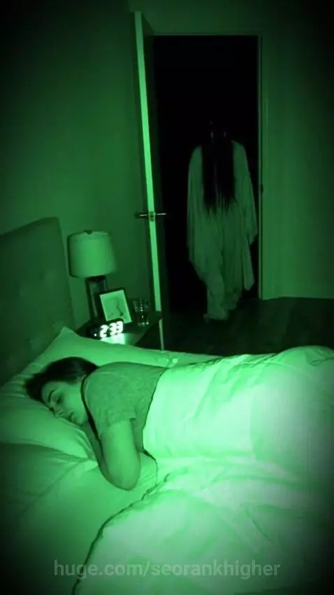 Night vision view of a dark figure with long hair lunging from a doorway towards a person sleeping in bed.