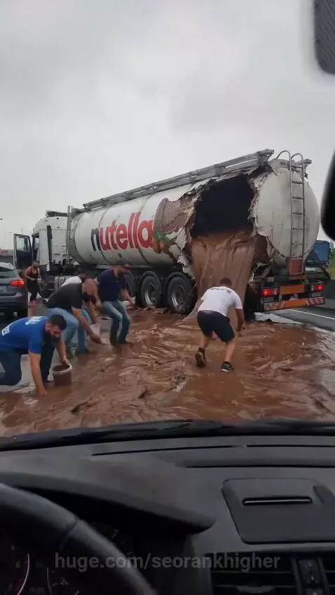 A damaged Nutella tanker truck spilling chocolate spread on a wet road, with people scooping it up.