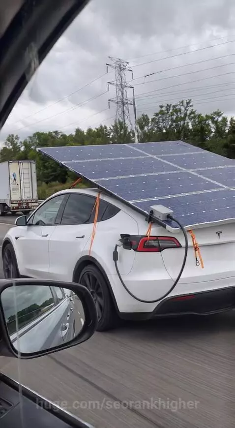 White Tesla car driving on highway with a large solar panel array attached to its rear, connected to the charging port.