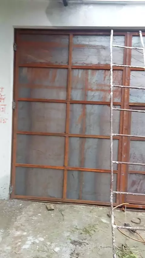 Rusty metal gate in an outdoor Indian workshop, with tools, a ladder, and red Hindi script on a white wall.