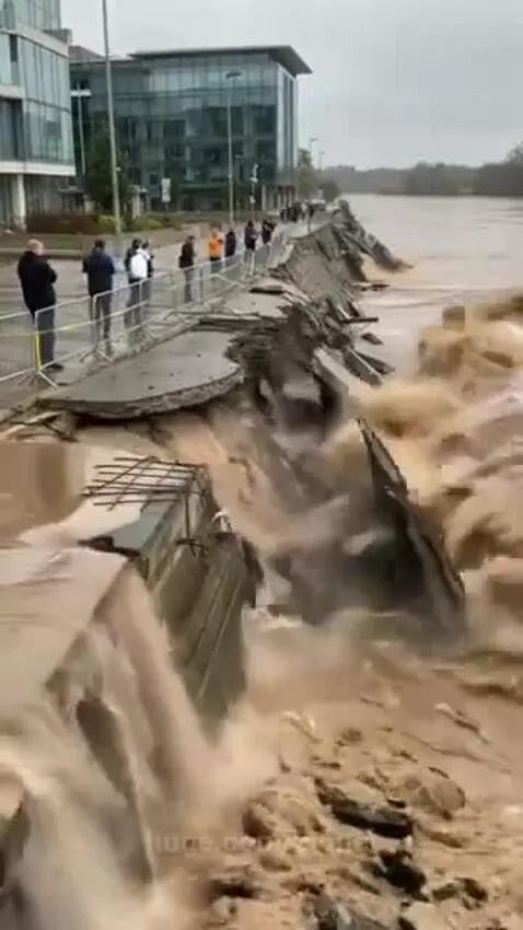 A large, muddy wave crashes against a concrete embankment, causing it to crumble and collapse.