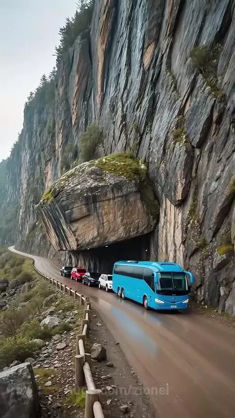 A large blue bus drives past a cliff face as rocks and dust fall towards the road.