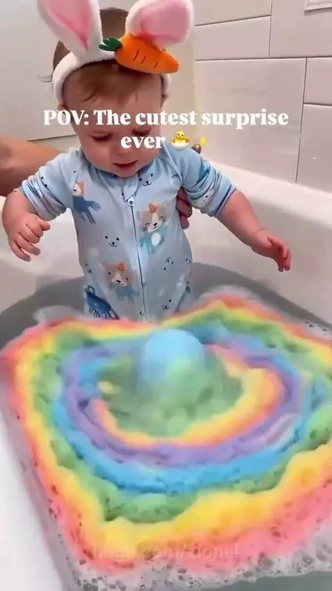 Baby in bunny ears smiling at a toy turtle emerging from rainbow bath bomb foam.