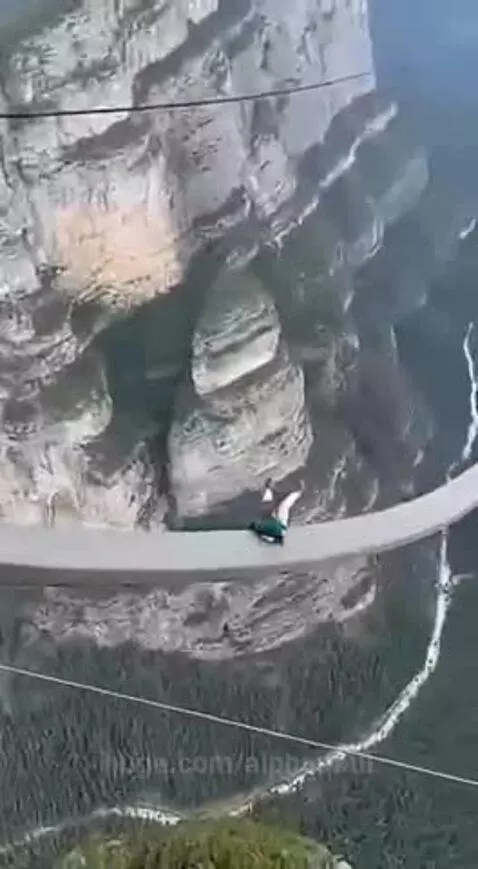 Woman in green jacket on a high cliff platform, about to slide down a long white track into a valley.