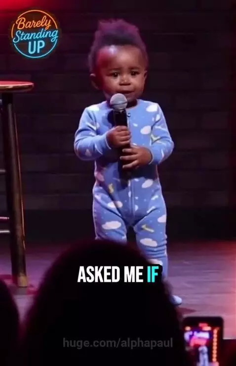 Baby with mohawk on stage holding microphone, dressed in a blue cloud onesie, performing stand-up comedy.