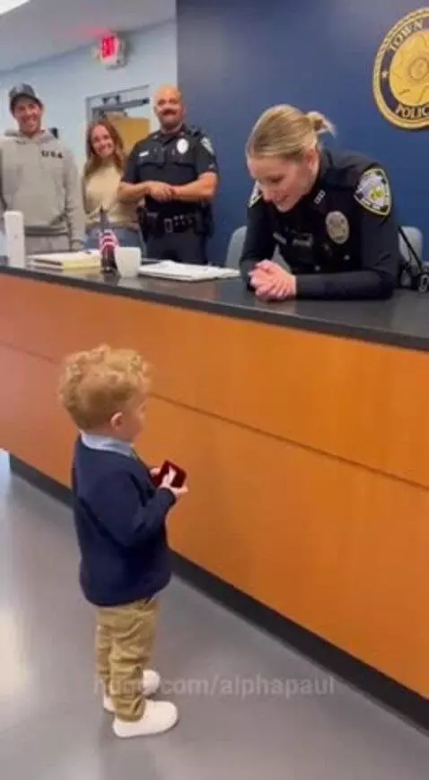 Young boy in blue sweater holds red ring box and proposes to a female police officer at a station counter.