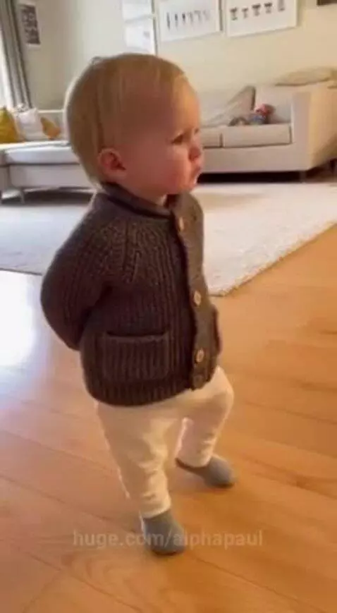 Blonde toddler in a grey cardigan standing on a wooden floor, looking at the camera.