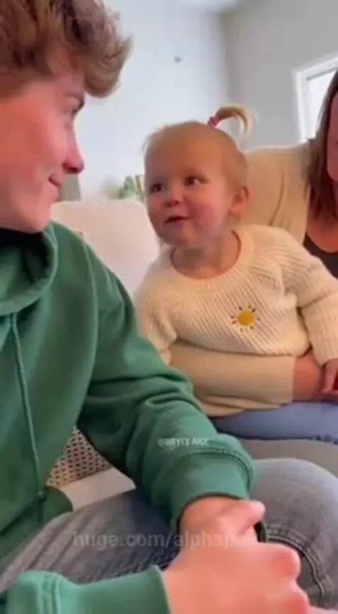 A blonde toddler in a sun sweater smiles at a man in a green hoodie on a couch.