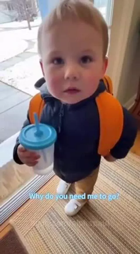 Young child in blue jacket and orange backpack holding a sippy cup, looking up and talking about a math test.