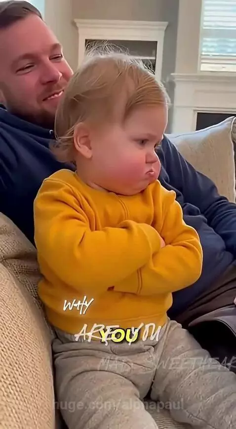 Blonde baby with arms crossed and a pouting expression on a couch, with a smiling man in the background.