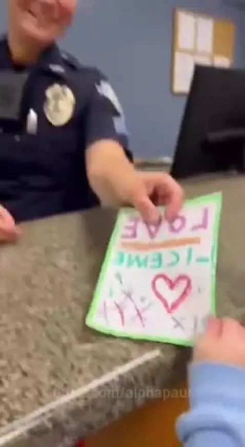 A young child in a blue hoodie hands a drawing labeled 'LOVE LICENSE' to a smiling female police officer.