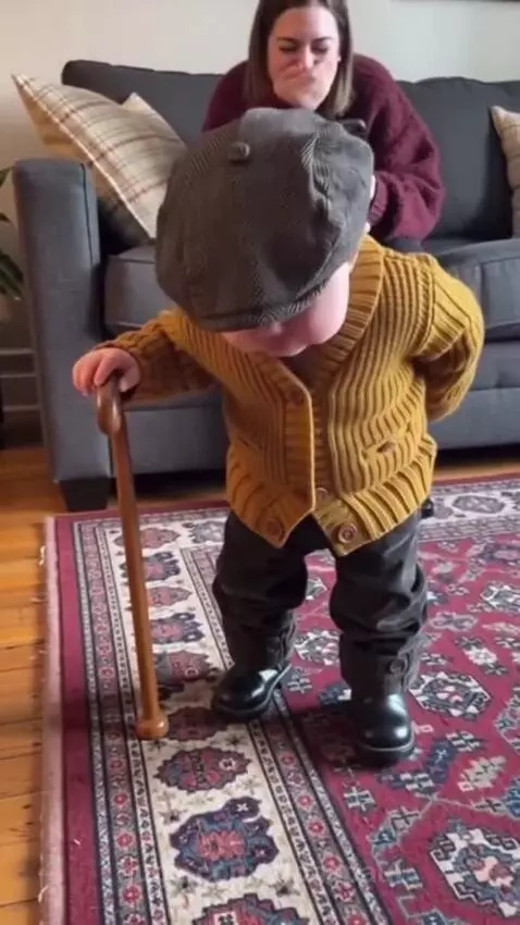 Baby dressed as a grandpa in a flat cap and cardigan, leaning on a walking stick and saying "My back hurts."