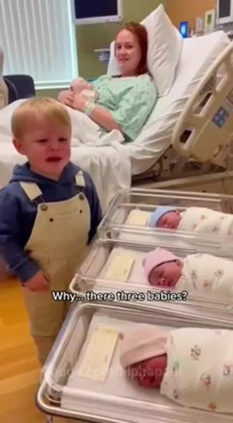 A crying young boy in overalls looks worriedly at three newborn babies in bassinets, while his mom holds another baby.