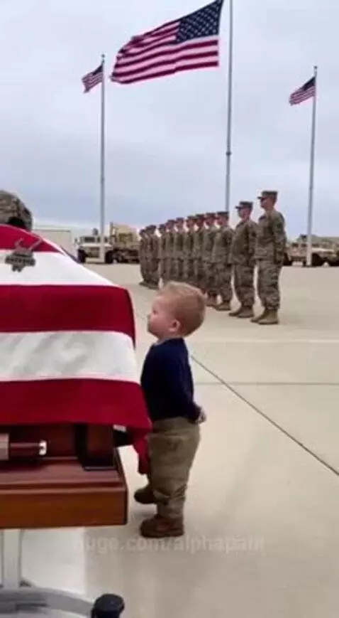 A toddler stands before a flag-draped coffin on an airfield, with military personnel in the background.