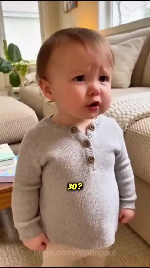 A baby with light brown hair in a grey shirt stands in a living room, looking off-camera and speaking.