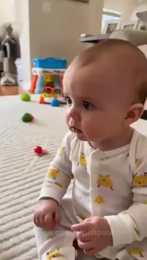 Baby in yellow chick pajamas crying as a cat swats its head on a rug.