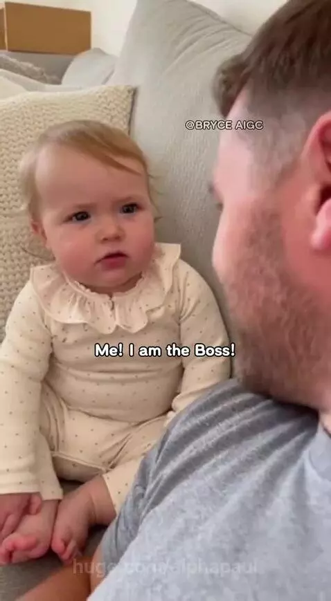 Baby girl in a polka-dotted onesie sitting on a couch, pointing at her dad and laughing.