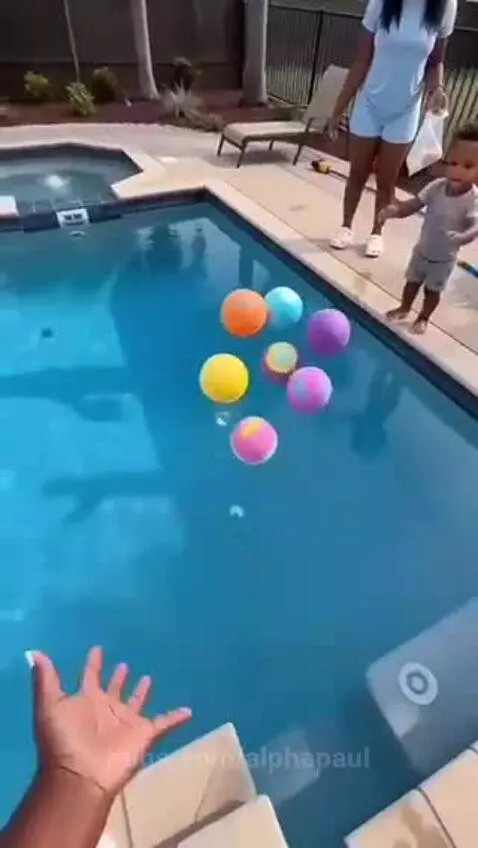 Child and adult watch as large, colorful bath bombs create vibrant foam in a swimming pool with toys.