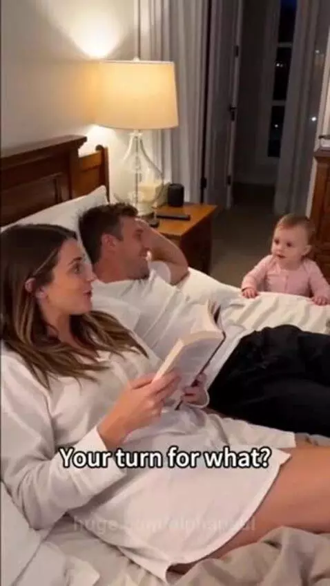 Baby sitting between parents in bed, looking at mom, while claiming 'my turn' with dad.
