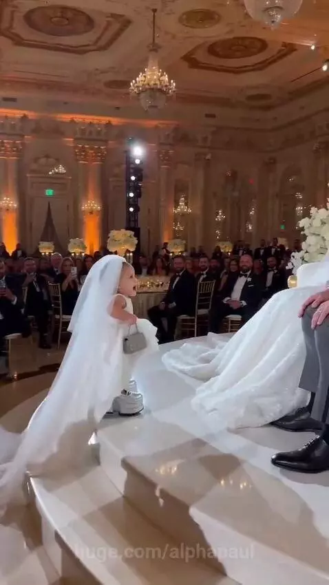 Child in a wedding dress and veil with oversized shoes approaches a bride and groom on a wedding reception stage.