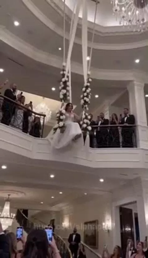 Bride in a white gown descending on a swing decorated with white flowers and ribbons, smiling at wedding guests.