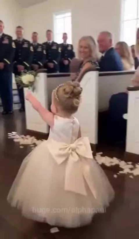 A young flower girl in a white dress and tiara scatters petals down a wedding aisle, waving to guests.