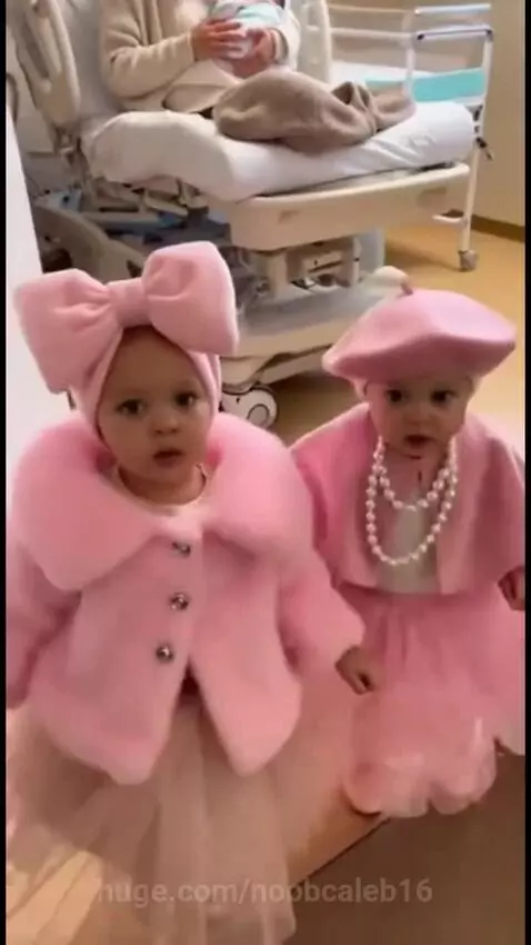 Two young babies in matching pink outfits walk hand-in-hand towards the camera in a hospital room, meeting their new baby sister.