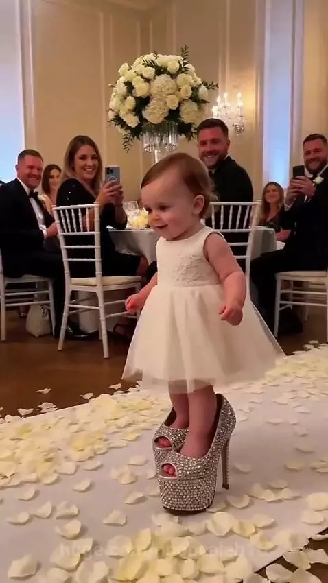 Baby dressed as a bride in oversized heels walks down a wedding aisle with rose petals.