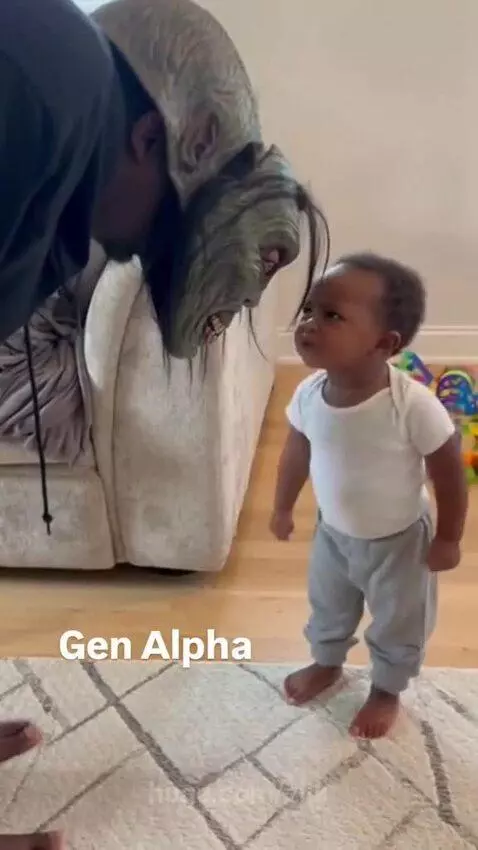 Baby in white t-shirt and grey pants scolding a person wearing a green monster mask.