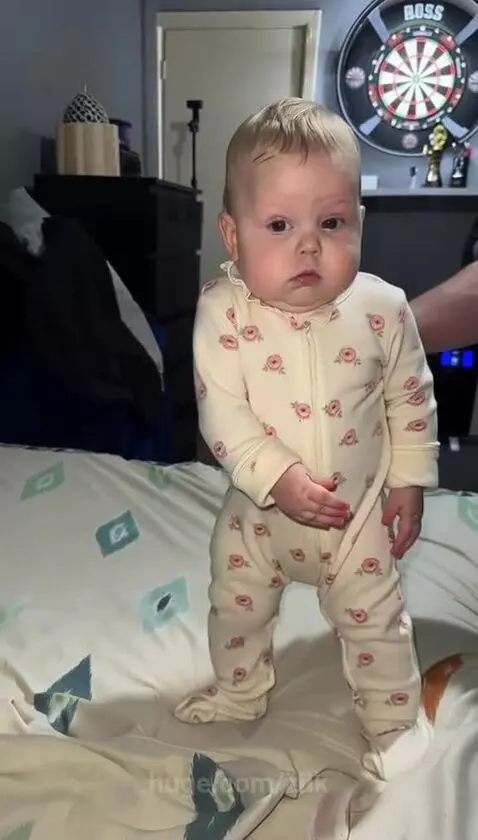 A baby in a floral sleeper stands on a bed, moving arms and legs rhythmically in a dance-like motion.