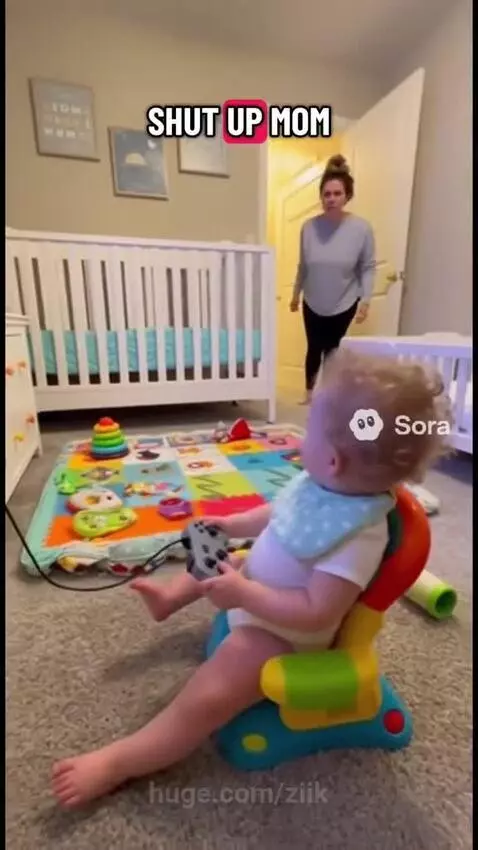 Baby with curly hair in a bib and diaper sits in a chair playing a video game, ignoring his surprised mother.
