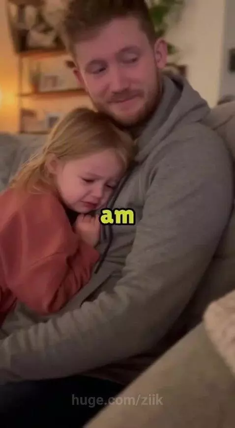 A young girl in a pink top crying and hugging her smiling father on a couch.