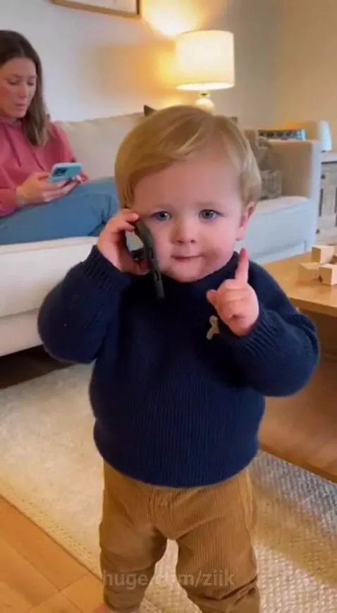 Toddler in a teddy bear sweater holding a phone to his ear, pretending to talk to his dad.