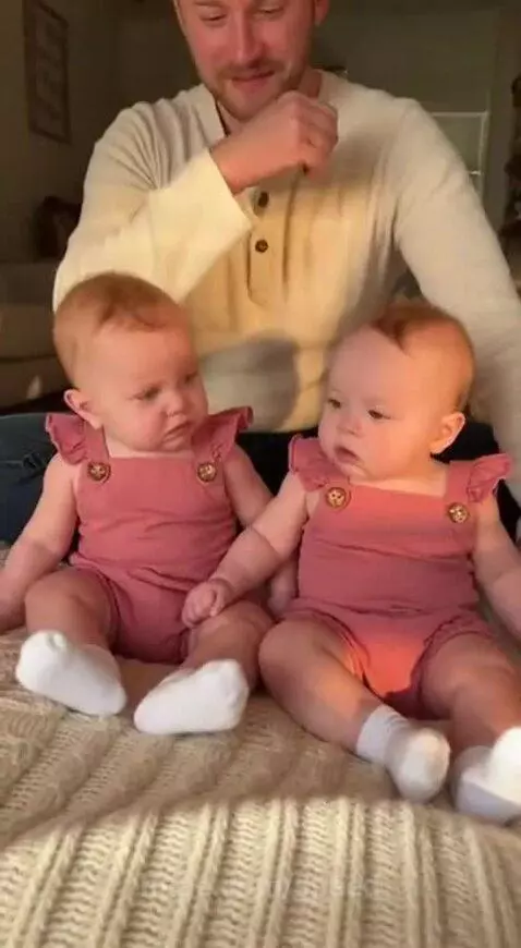 Two babies in pink rompers and white socks sit with a man on a bed, one baby attempts to mimic a sneeze.