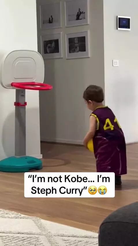Young child in a purple and yellow jersey shoots a basketball into a small hoop, smiling.