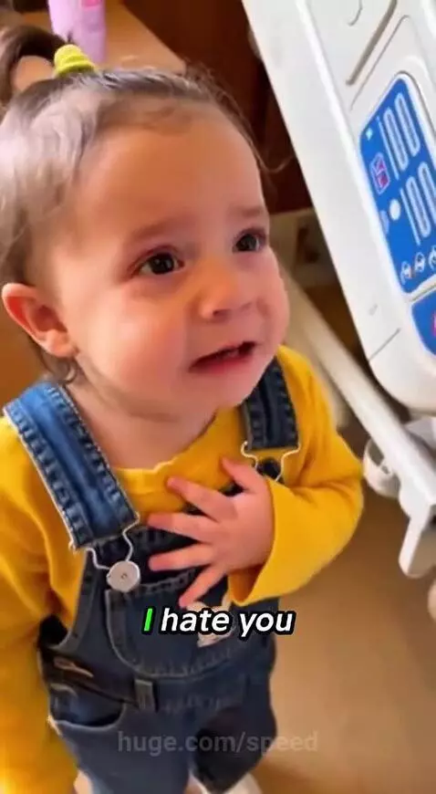 Toddler looking upset and crossing arms next to parents holding a newborn baby in a hospital bed.