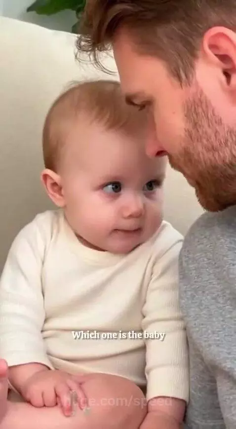 Close-up of a baby with blue eyes looking at a bearded man, asking why mom calls him 'baby.'