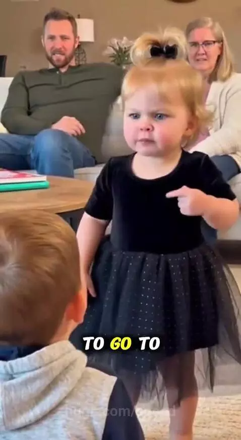Young girl with blonde hair and a bow, wearing a black dress, scolding someone with an angry face and pointing finger.