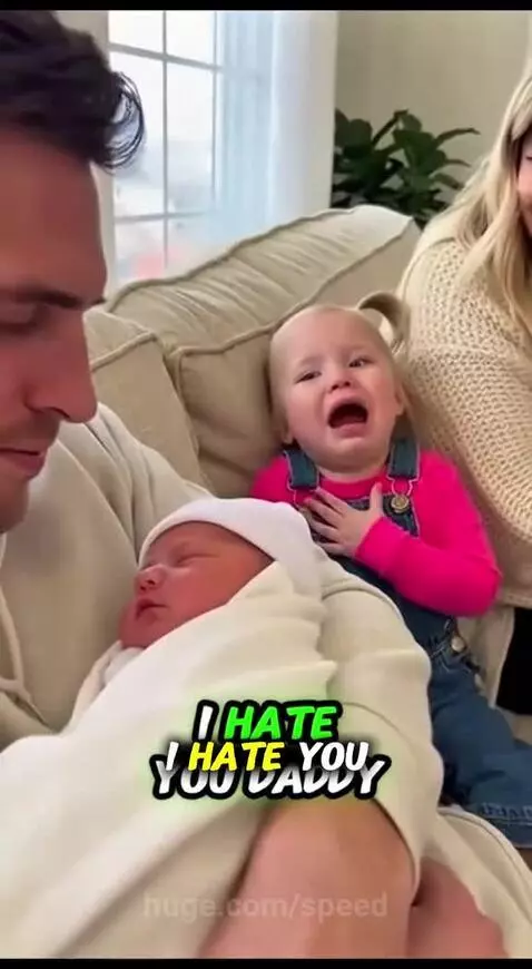 Upset little girl with pigtails crying next to a newborn baby held by her dad.