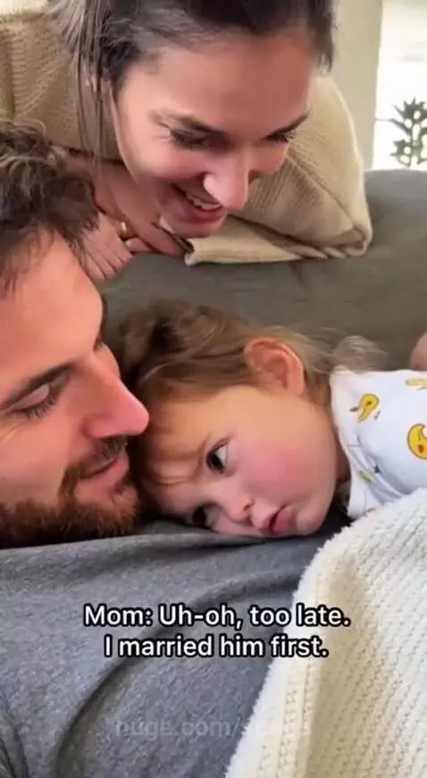 A young girl rests her head on her dad's chest while he lies down; mom smiles behind them.