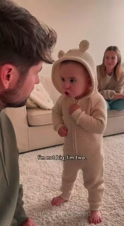 A stern toddler in a bear-eared onesie stands barefoot, looking at a kneeling man, while a woman smiles on the couch.