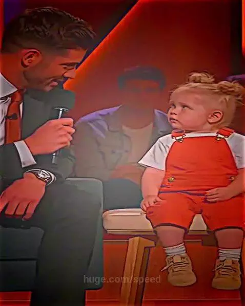 A young child in orange overalls and hair buns on a stage, looking serious while explaining her dad's reaction to her mom's anger.