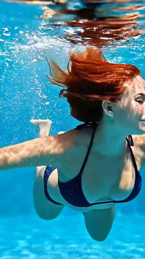 Woman with red hair in a blue bikini smiles underwater in a swimming pool, with city buildings visible in the background.