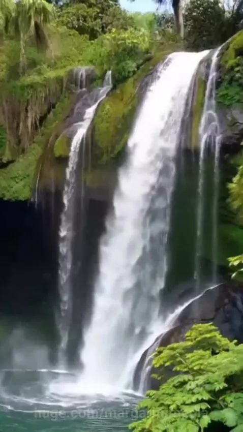 Aerial view of a tropical waterfall cascading onto a white sand beach with turquoise ocean and lush green mountains.