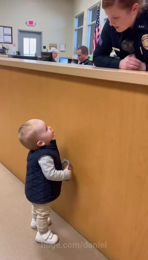 Baby in vest and hat holds toy handcuffs, looking up at smiling female police officer.