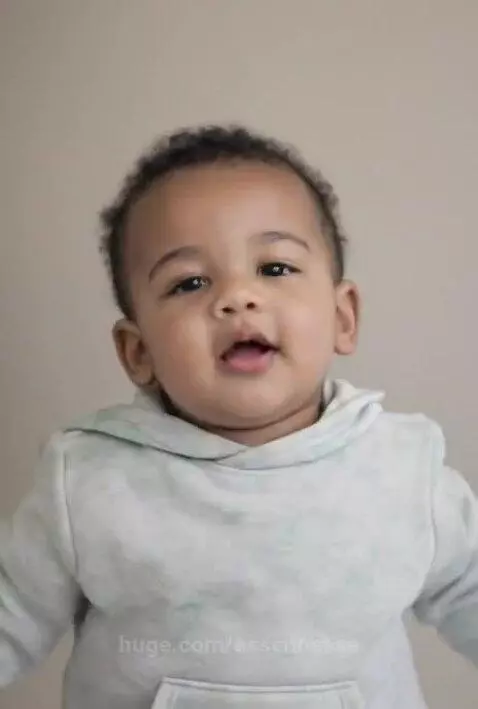Close-up of a baby with dark curly hair and light brown skin smiling and clapping its arms up and down.