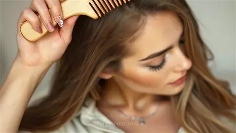 Young woman with long brown hair brushing it slowly with a wooden comb, eyes closed in relaxation.